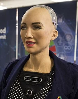 robot with a human-like face, wearing a dark jacket, displaying a friendly expression in a tech environment