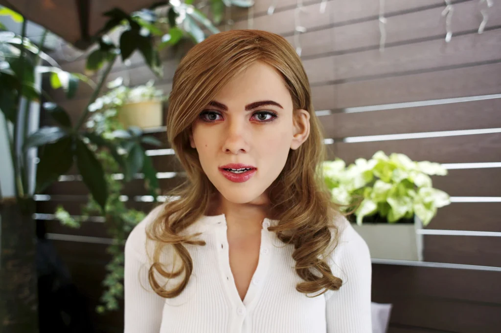 Realistic humanoid robot with long hair, wearing a white top, surrounded by greenery in a modern setting.